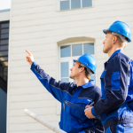 Deux techniciens en uniforme bleu et casque de sécurité observent la façade d’un bâtiment.