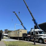 Deux grues soulèvent un camp forestier modulaire près d’un camion-remorque.