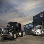 Camion Volvo noir stationné devant le bâtiment de Volvo Lac-St-Jean, avec un camion blanc à droite.
